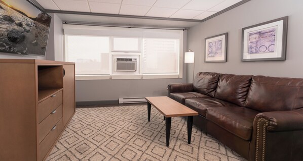 A room with a brown leather sofa, a wooden table, a brown cabinet, a patterned carpet, a window with an air conditioner, and two framed pictures on the wall. - Prestige Oceanview Hotel Prince Rupert (Prince Rupert)