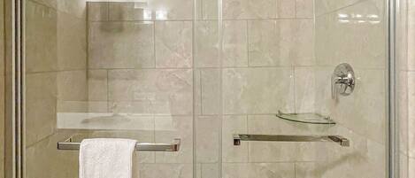 A glass-enclosed shower stall with a white towel hanging on a rack, a handheld showerhead, and a small shelf with toiletries.