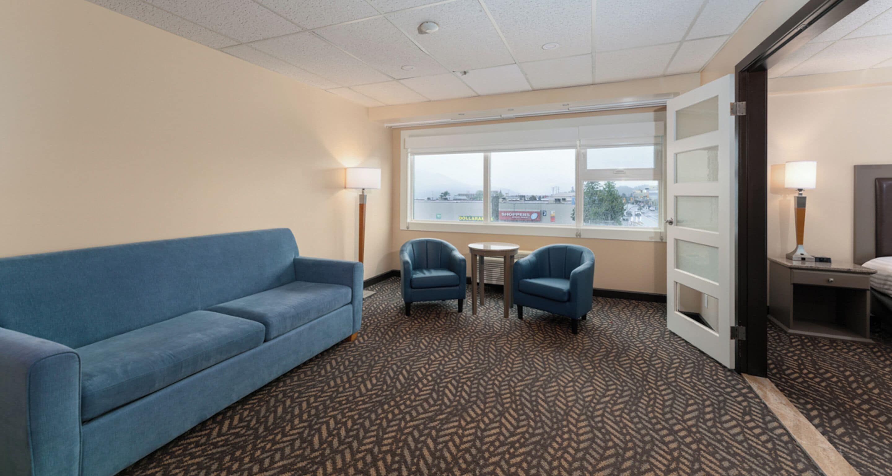A hotel room with a blue sofa, two armchairs, a small table, a lamp, a TV, and a window with a view of the city.