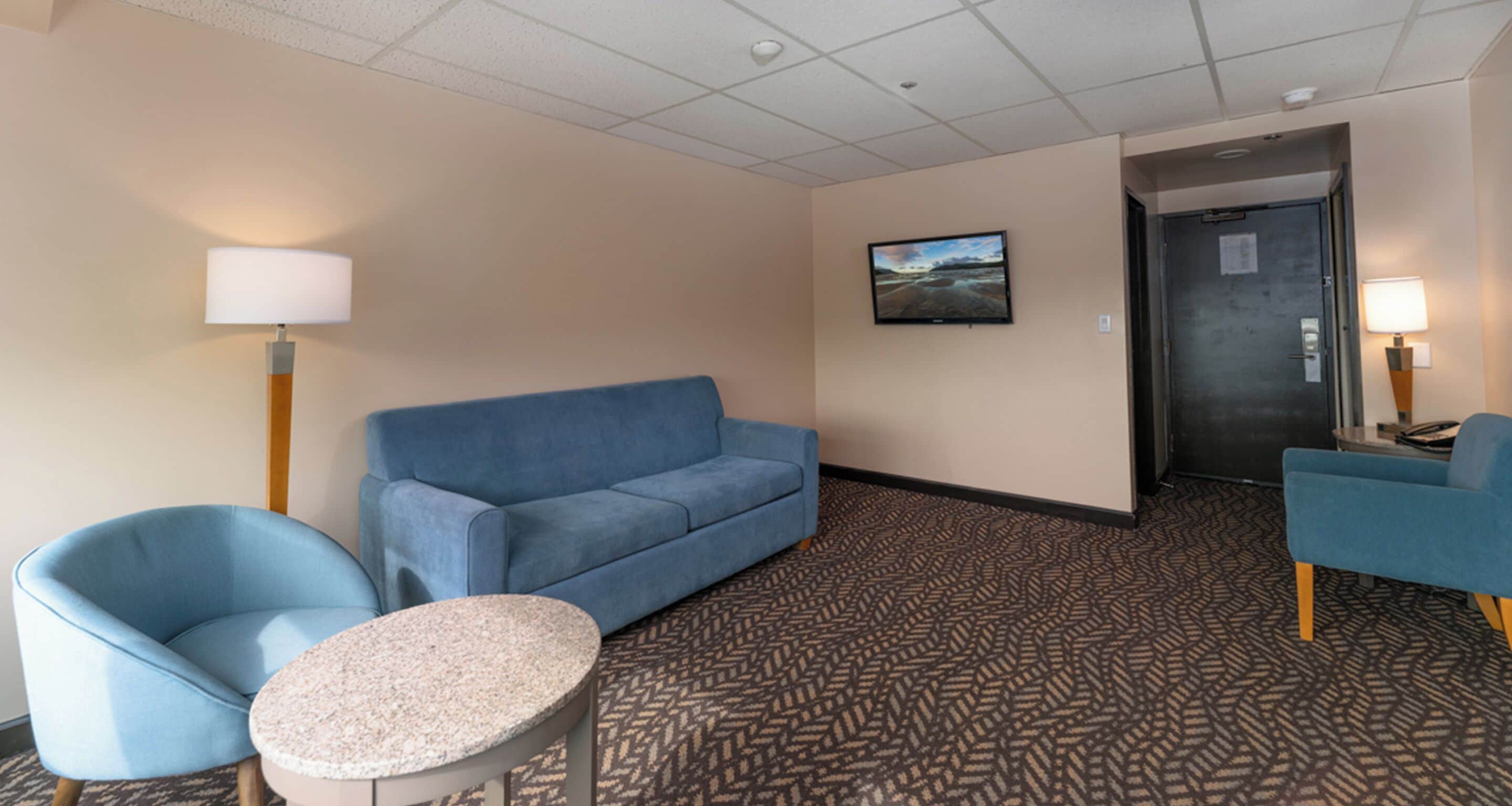 A hotel room with a blue sofa, two blue armchairs, a small round table, a television mounted on the wall, and a patterned carpet.