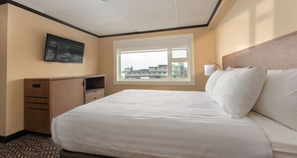 A hotel room with a large bed, a television on the wall, a window with a view of buildings, and a desk with drawers. - Prestige Oceanview Hotel Prince Rupert (Prince Rupert)
