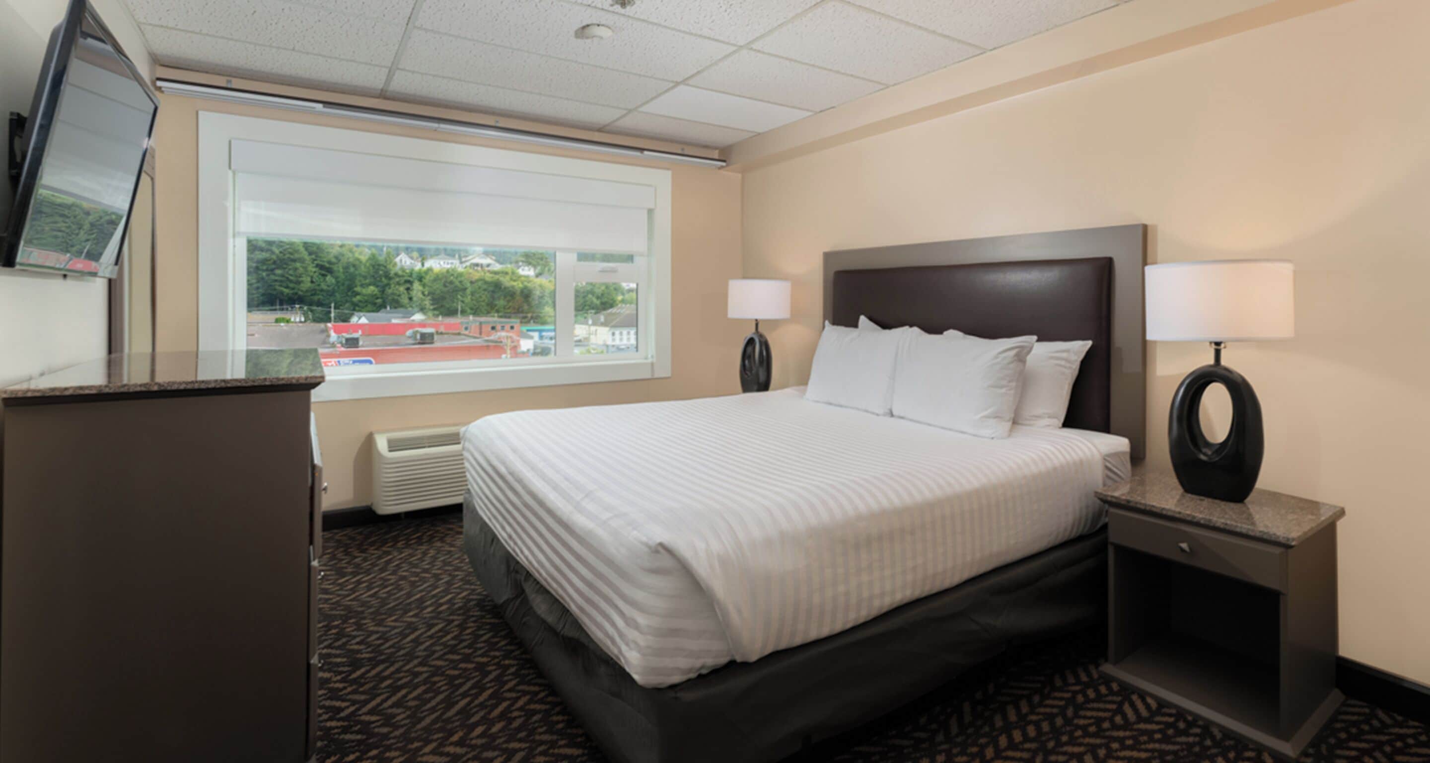 Habitación de hotel con una cama grande, dos lámparas de noche, un escritorio con televisor y una ventana con vistas a edificios y árboles.