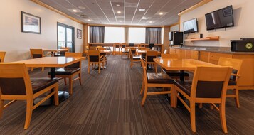 A hotel dining area with multiple tables and chairs, a television on the wall, and a kitchen counter.