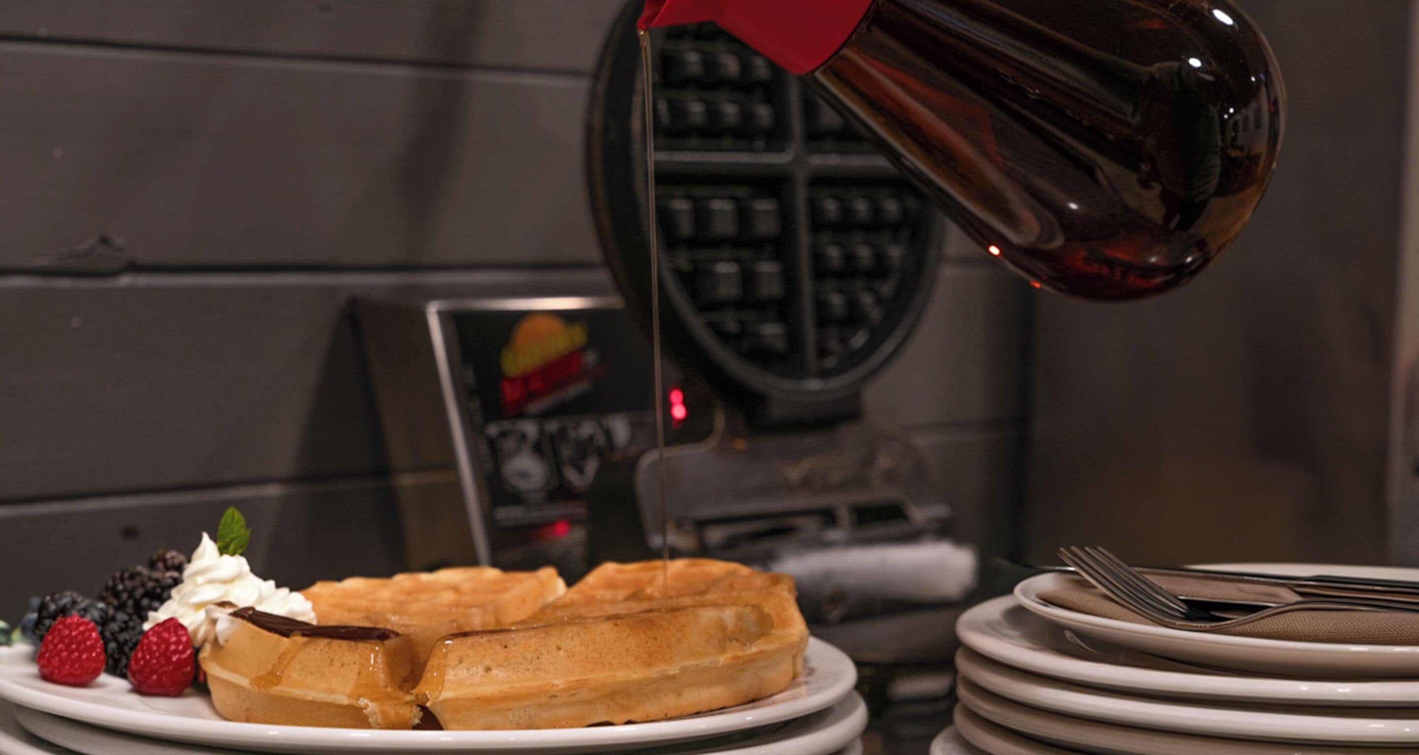 A waffle with whipped cream and berries on a plate, a stack of plates, and a waffle iron in the background.