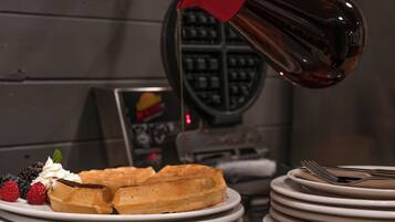 A waffle with whipped cream and berries on a plate, a stack of plates, and a waffle iron in the background.