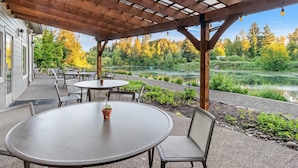 Un patio al aire libre con mesas y sillas redondas, una pérgola de madera y vistas a un lago y un bosque.
