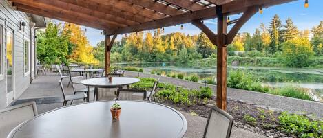 Un patio al aire libre con mesas y sillas redondas, una pérgola de madera y vistas a un lago y un bosque.