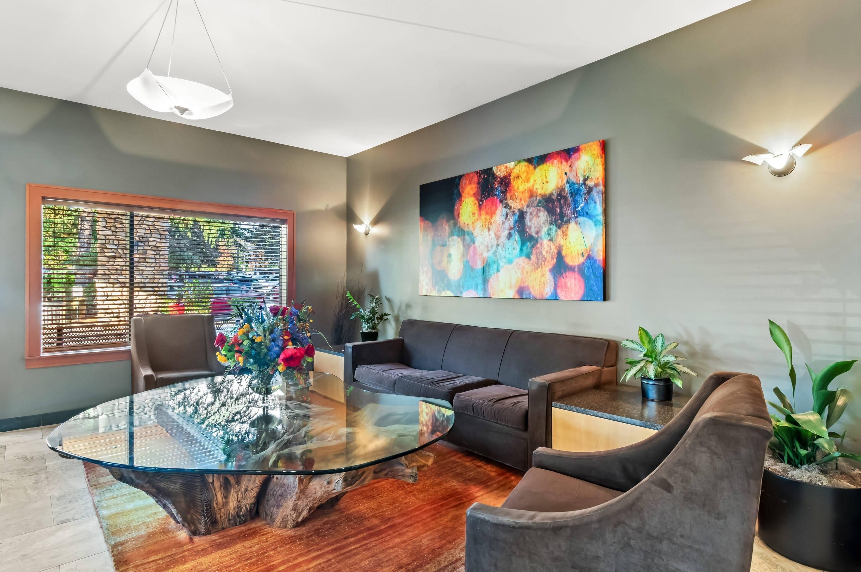 A modern living room with a glass coffee table, leather sofas, a large abstract painting, and a window with a view of parked cars.