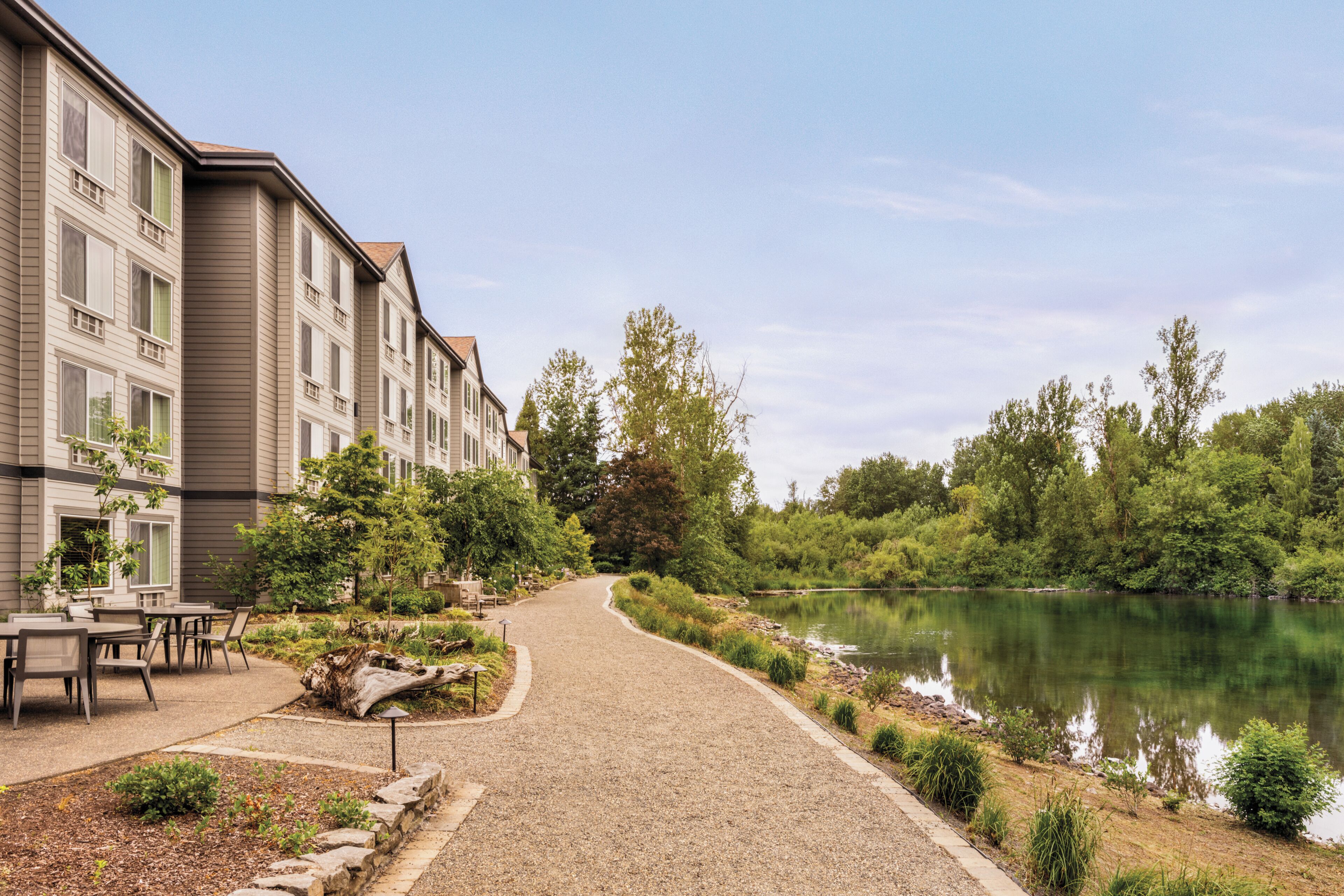 A pathway leading to a building with multiple windows, surrounded by greenery and a body of water.