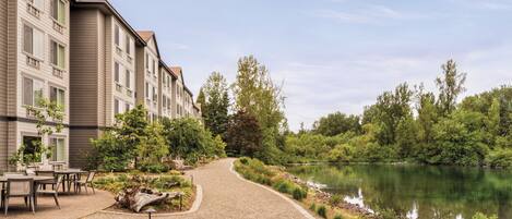 A pathway leading to a building with multiple windows, surrounded by greenery and a body of water.