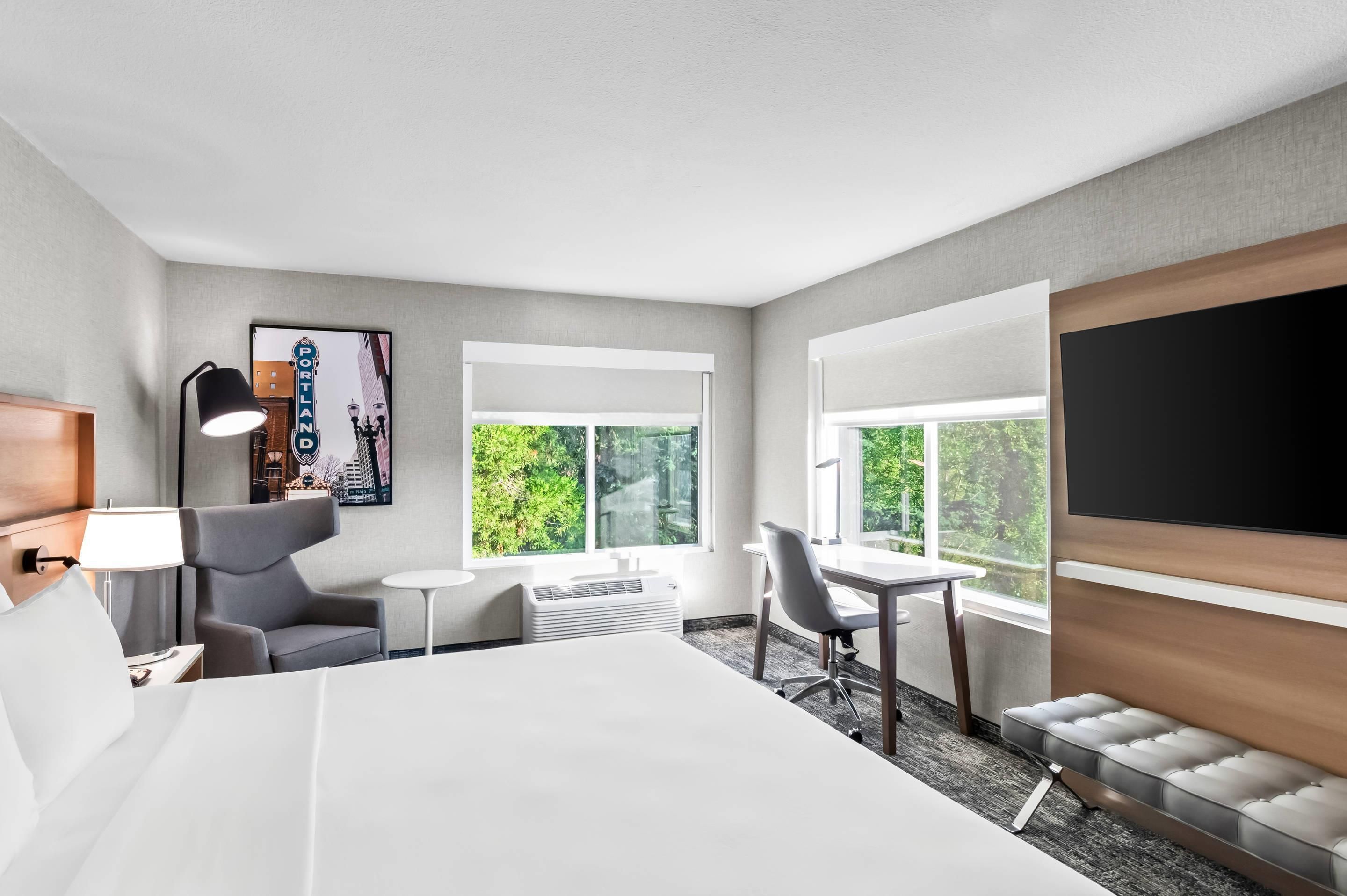 A hotel room with a bed, a desk, a chair, a TV, and a window with a view of greenery.