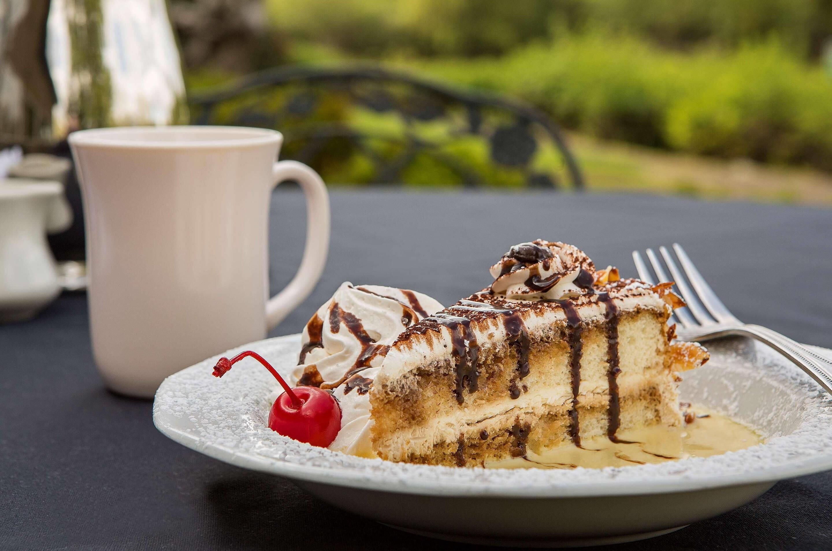 Una fetta di torta con panna montata e salsa al cioccolato su un piatto, insieme a una ciliegina e una tazza bianca, su un tavolo all'aperto.