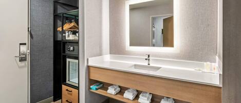A modern bathroom with a large mirror, white sink, and wooden vanity. There is a storage closet with hanging clothes and a shoe rack.