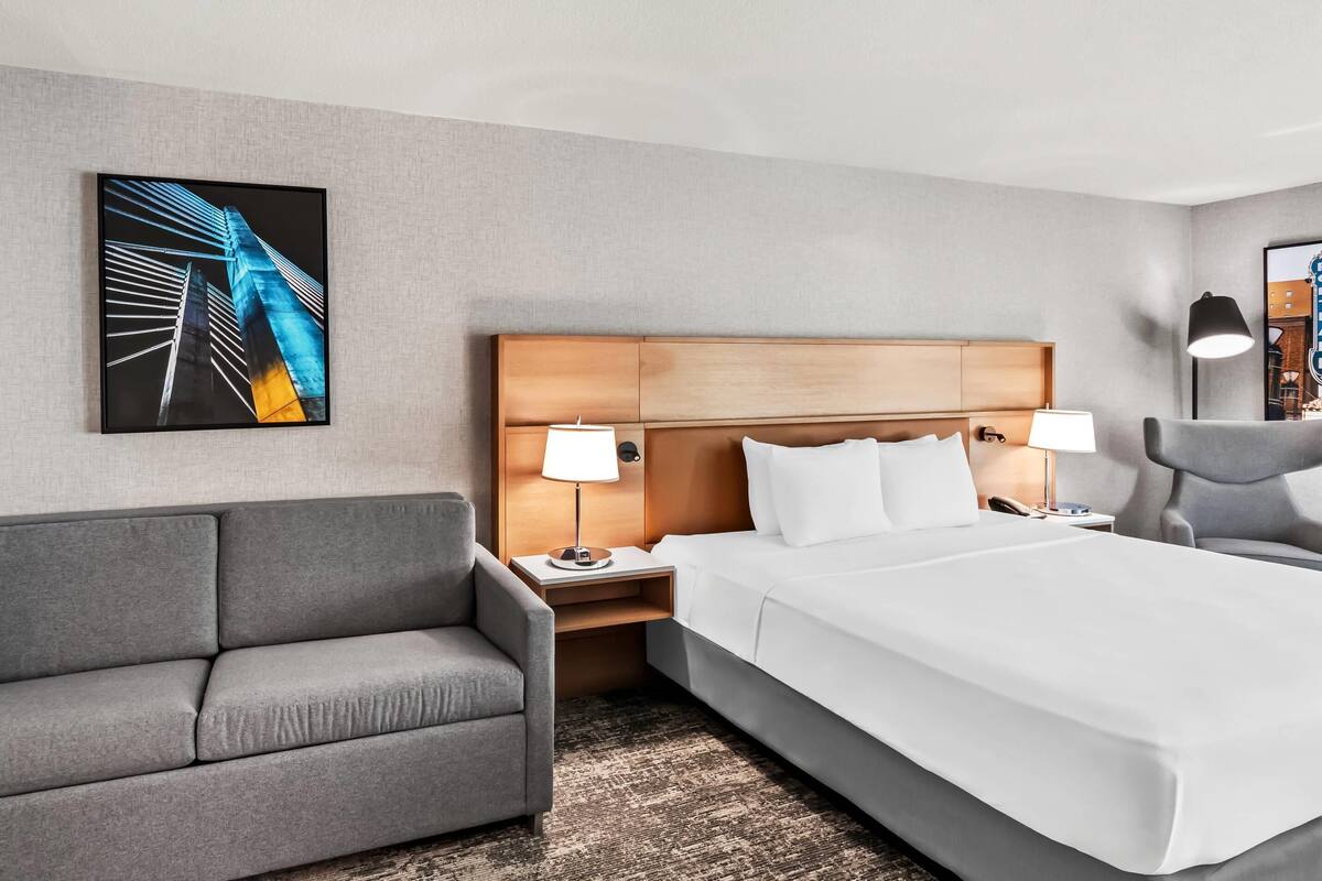 A hotel room with a large bed, a gray sofa, two bedside tables with lamps, and a framed picture on the wall.