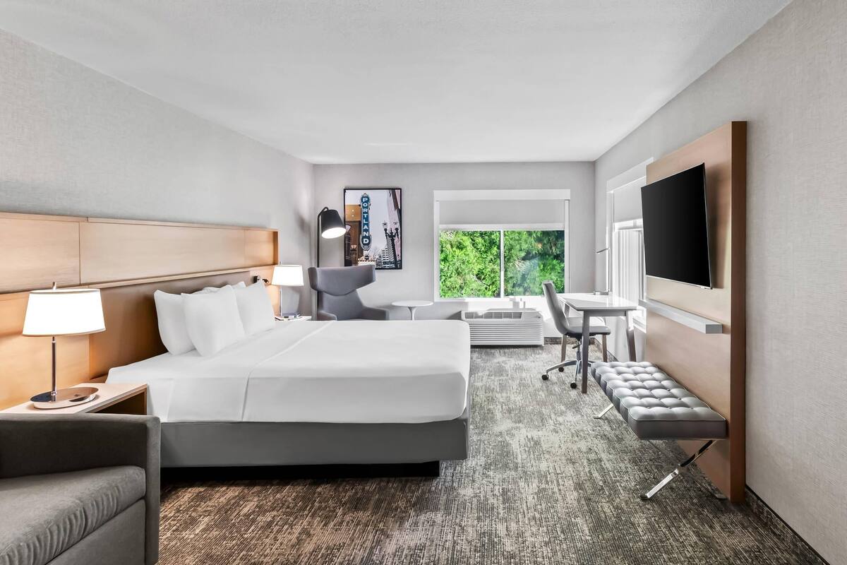 A hotel room with a bed, bedside table, lamp, chair, desk, and TV.