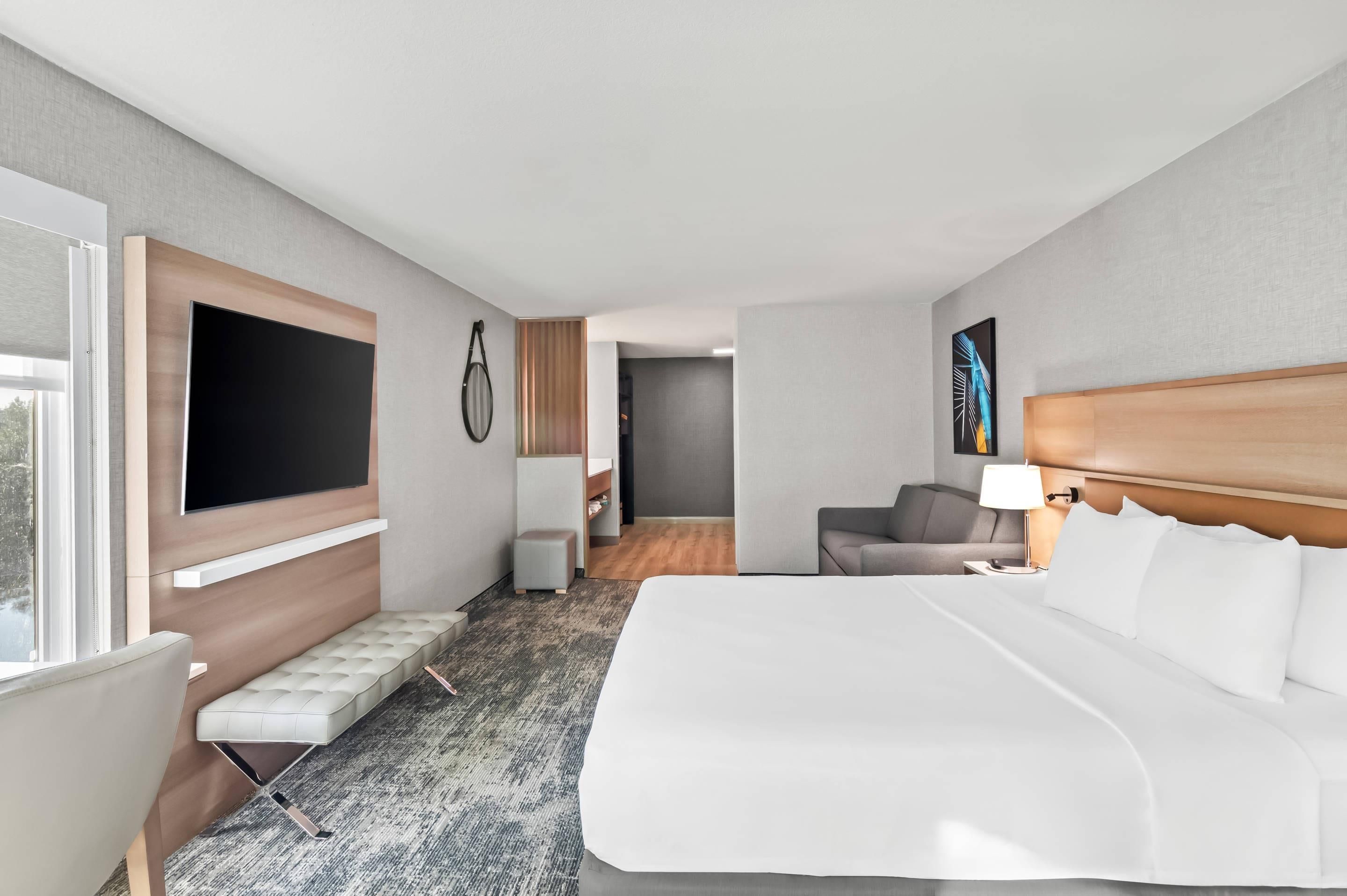 A modern hotel room with a large bed, a seating area with a bench, a flat-screen TV mounted on the wall, a desk with a lamp, and a view of the hallway.