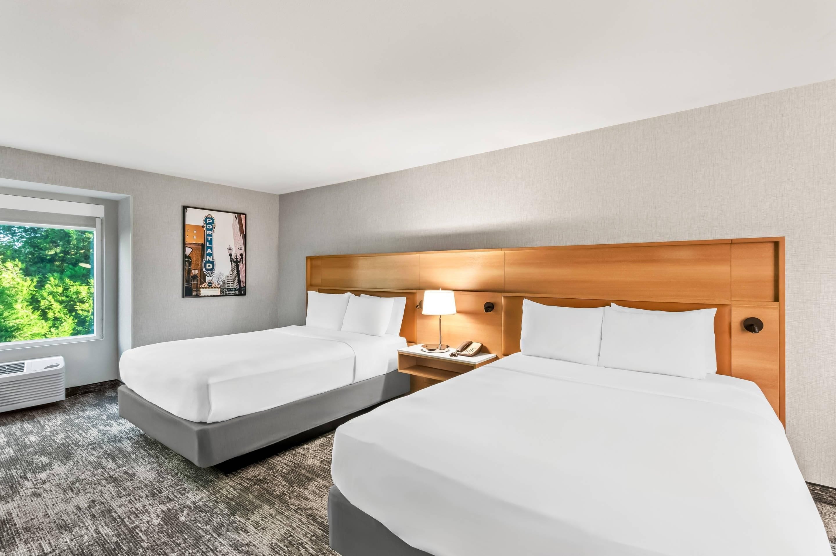 Habitación de hotel con dos camas, cabecero de madera, mesita de noche con lámpara y ventana con vista a zonas verdes.