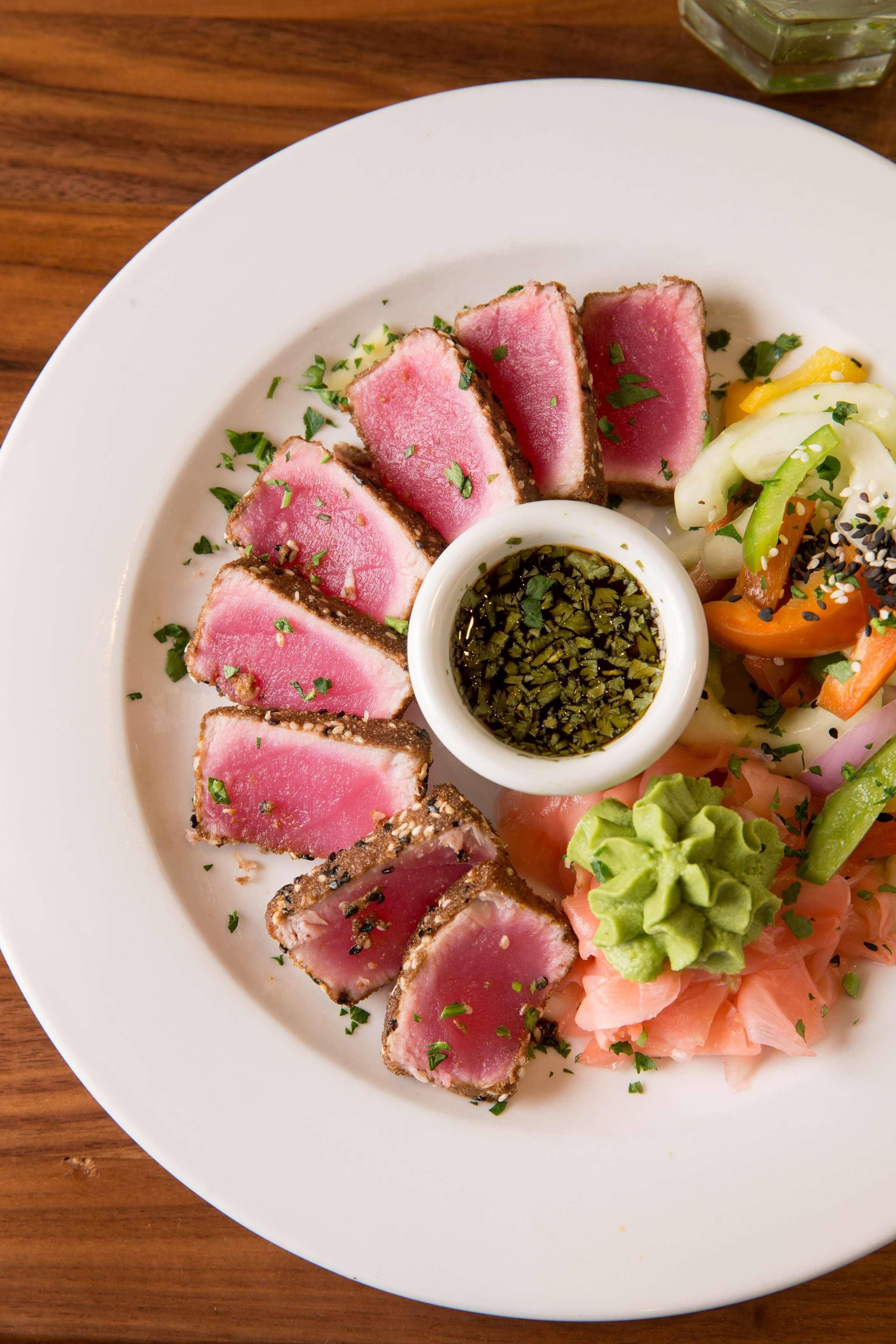 A plate of sliced tuna, avocado, and vegetables with a small bowl of sauce.