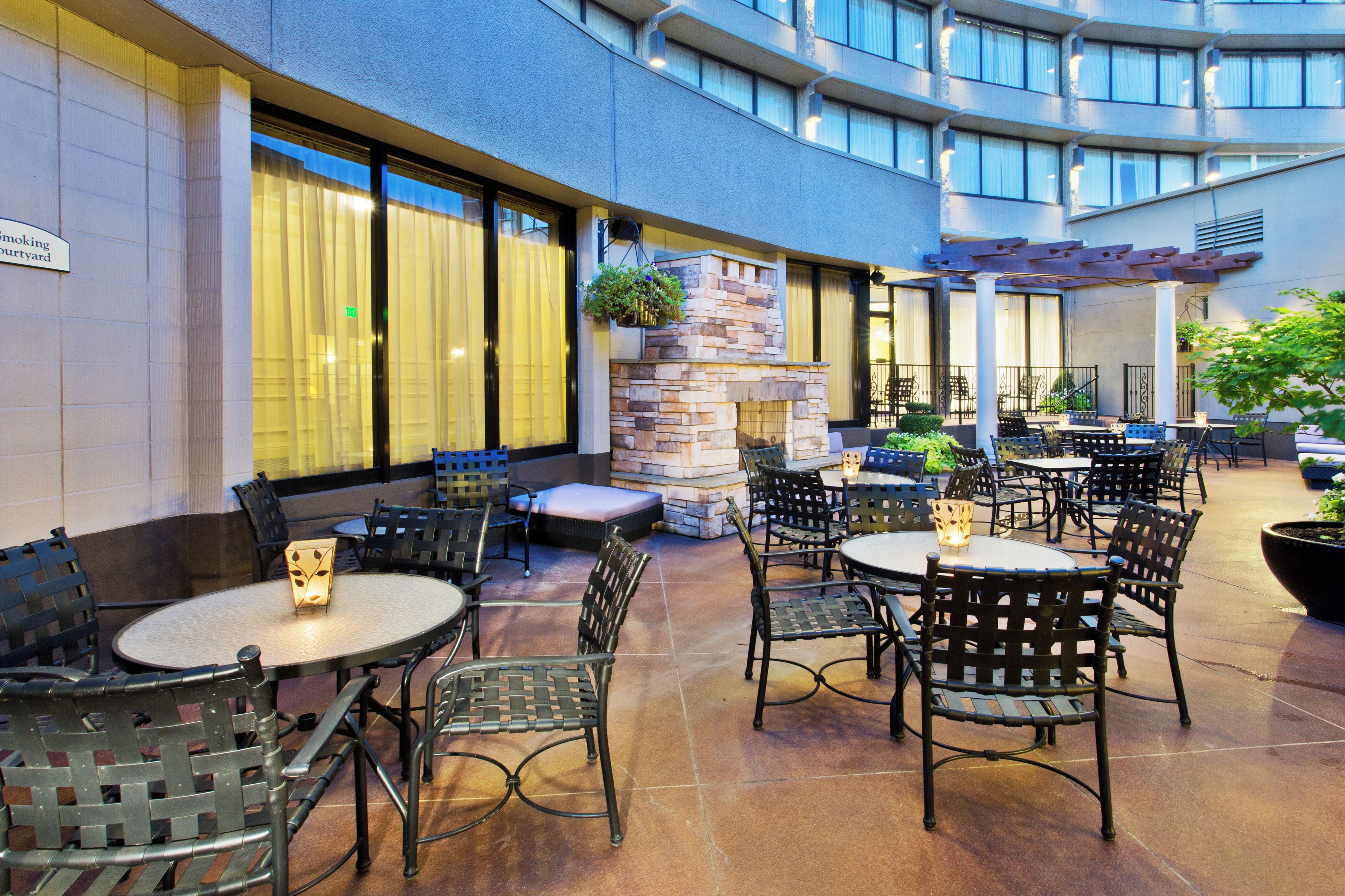 An outdoor patio area with round tables and chairs, a stone fireplace, and a view of the building's interior through large windows.