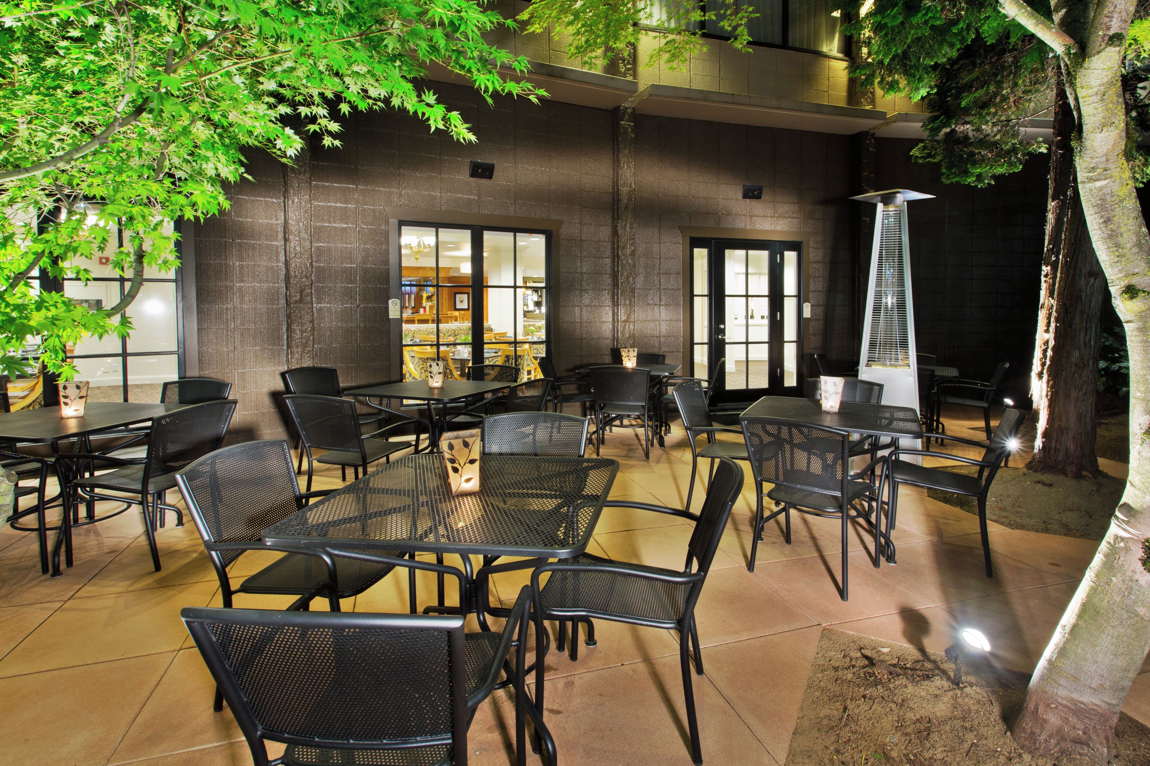 An outdoor dining area with tables and chairs, a building with large windows, and a tree.