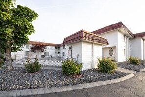 A white building with a red roof, surrounded by a gravel area and small bushes.