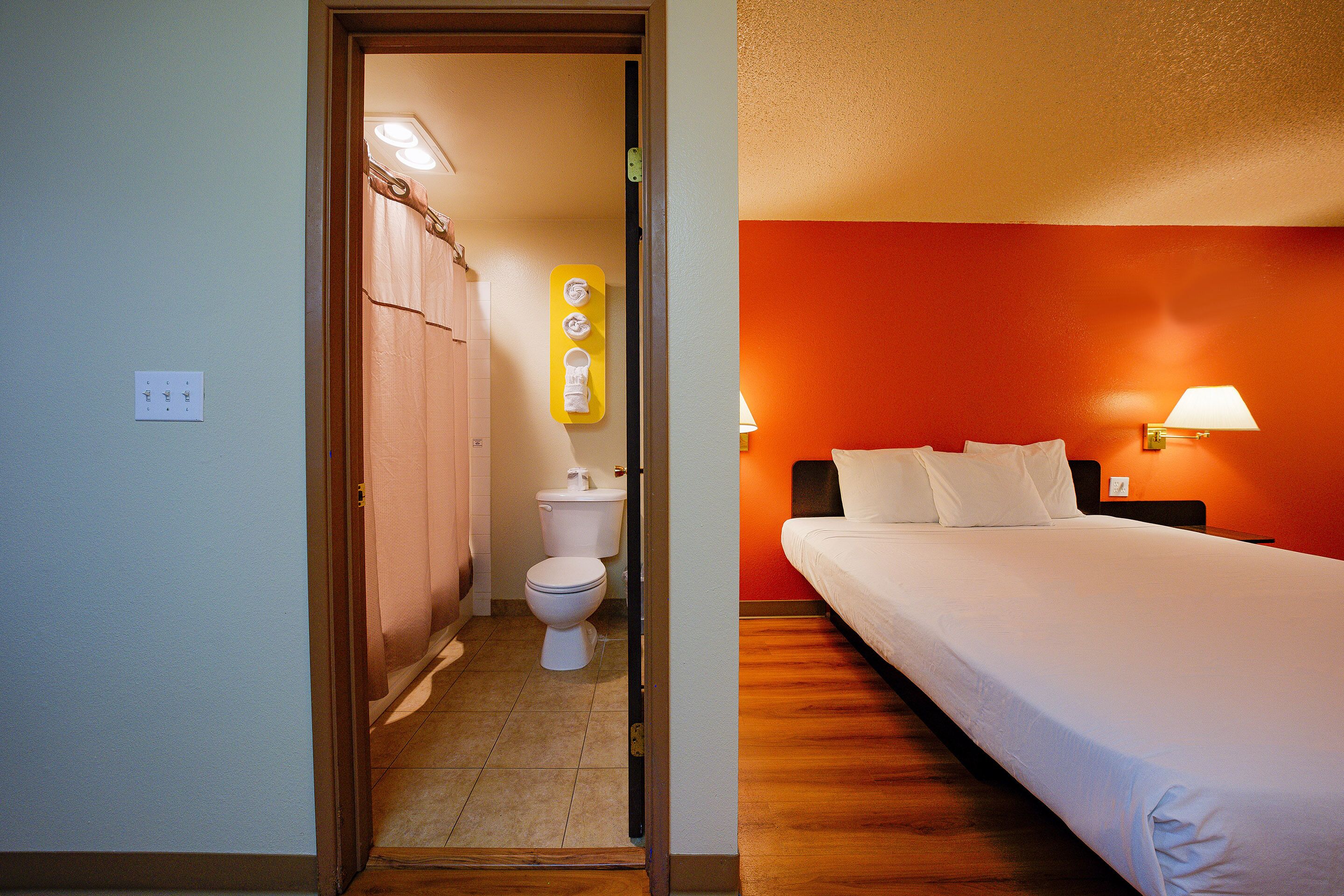 A hotel room with a wooden floor, a bed with white bedding, a nightstand with a lamp, and a bathroom with a toilet and shower curtain.