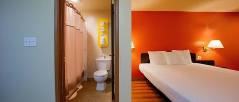 A hotel room with a wooden floor, a bed with white bedding, a nightstand with a lamp, and a bathroom with a toilet and shower curtain.