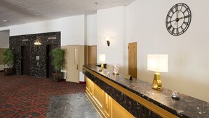 A hotel reception area with a marble counter, two potted plants, a clock, and a red carpeted floor. - Hôtels Gouverneur Montréal - Île Charron (Longueuil)