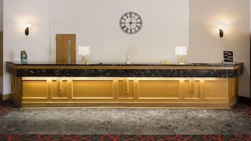 A reception desk with a marble top, gold paneling, two wall-mounted lamps, a clock, and a sign with room names.