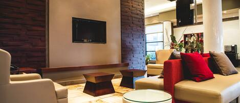 Ein modernes Wohnzimmer mit Sofa, Sessel und Couchtisch. An der Wand ist ein Flachbildfernseher angebracht und in der Ecke steht eine Zierpflanze.