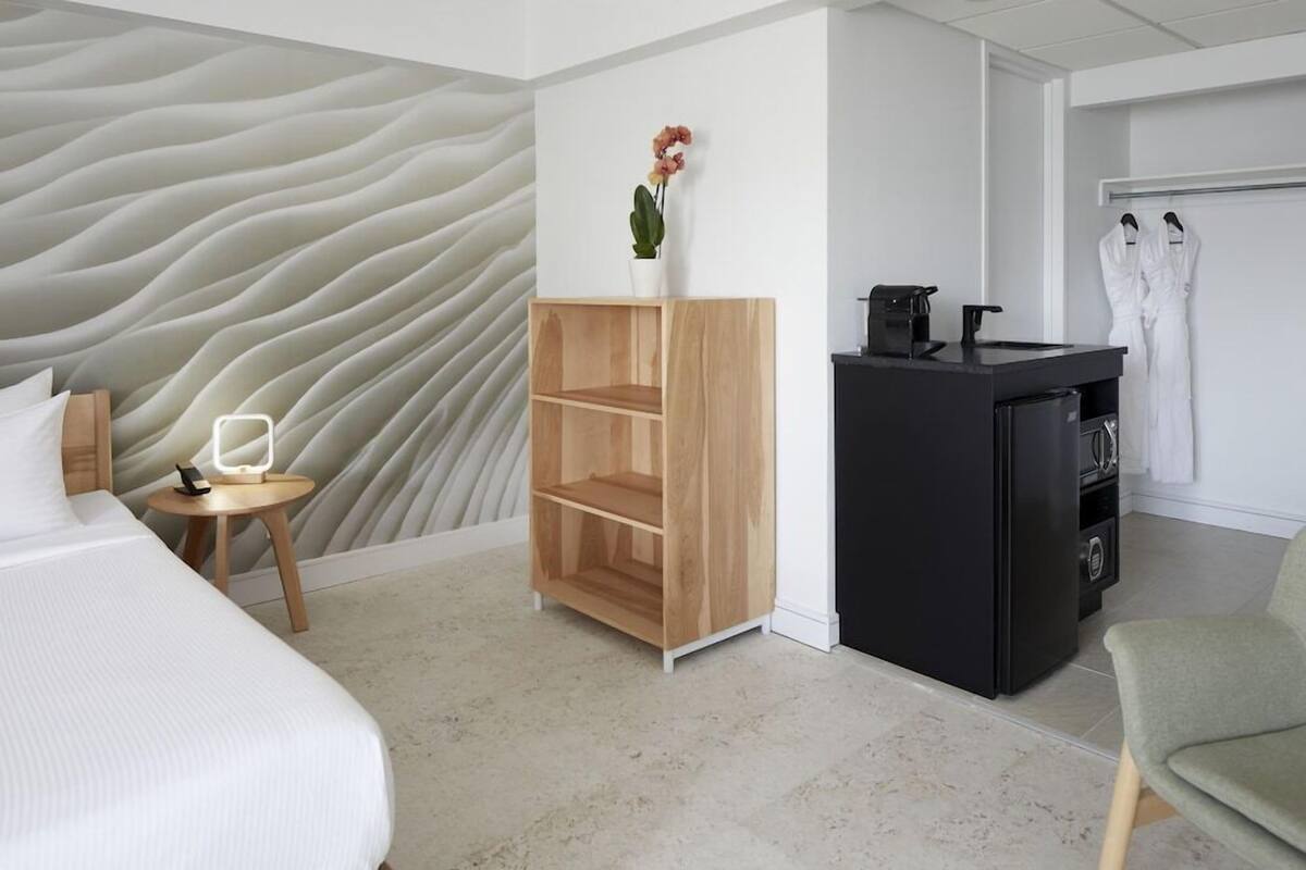 A hotel room with a bed, a small wooden shelf with a plant, a black minibar, and a robe rack.
