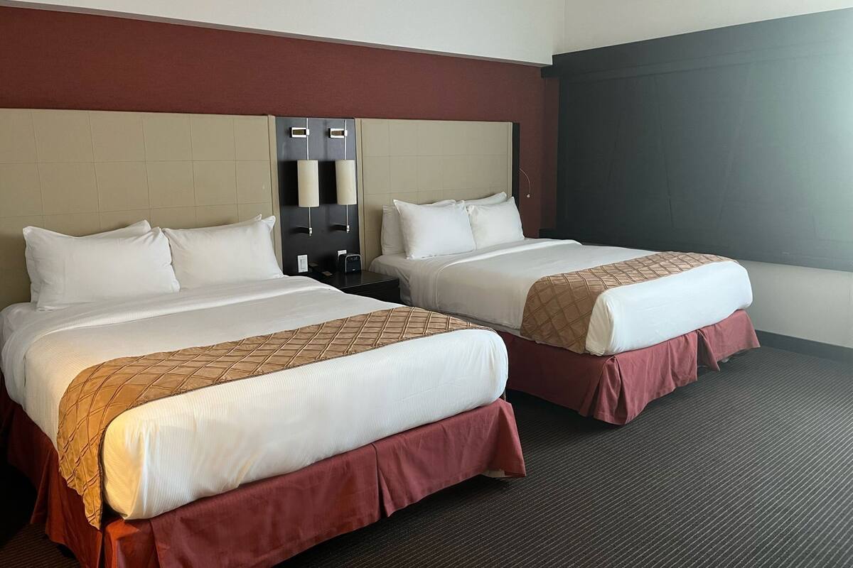 A hotel room with two beds, a dark carpet, a large window, and a wall-mounted light fixture.