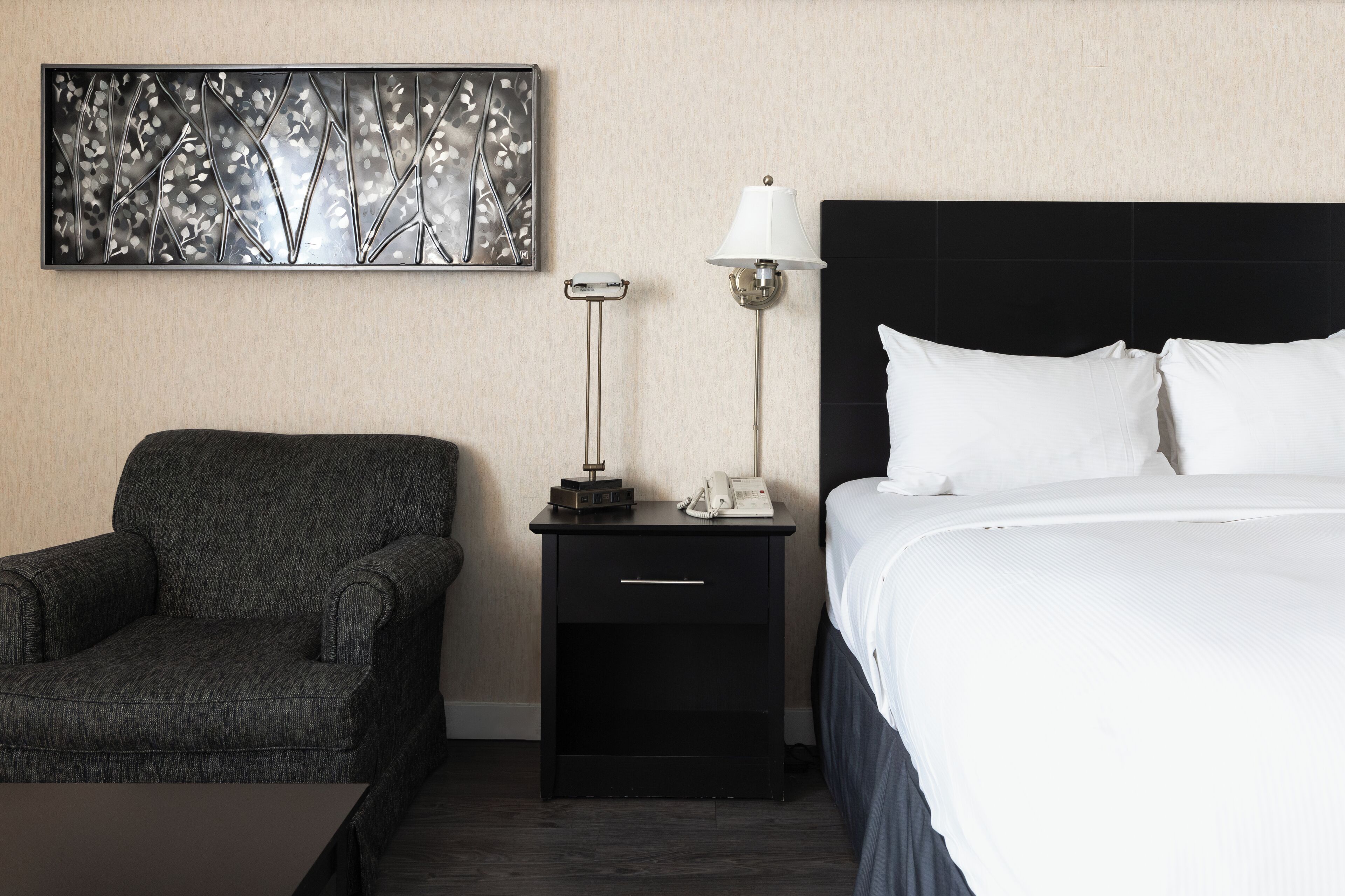 A hotel room with a bed, a nightstand with a lamp, a chair, and a decorative wall piece.