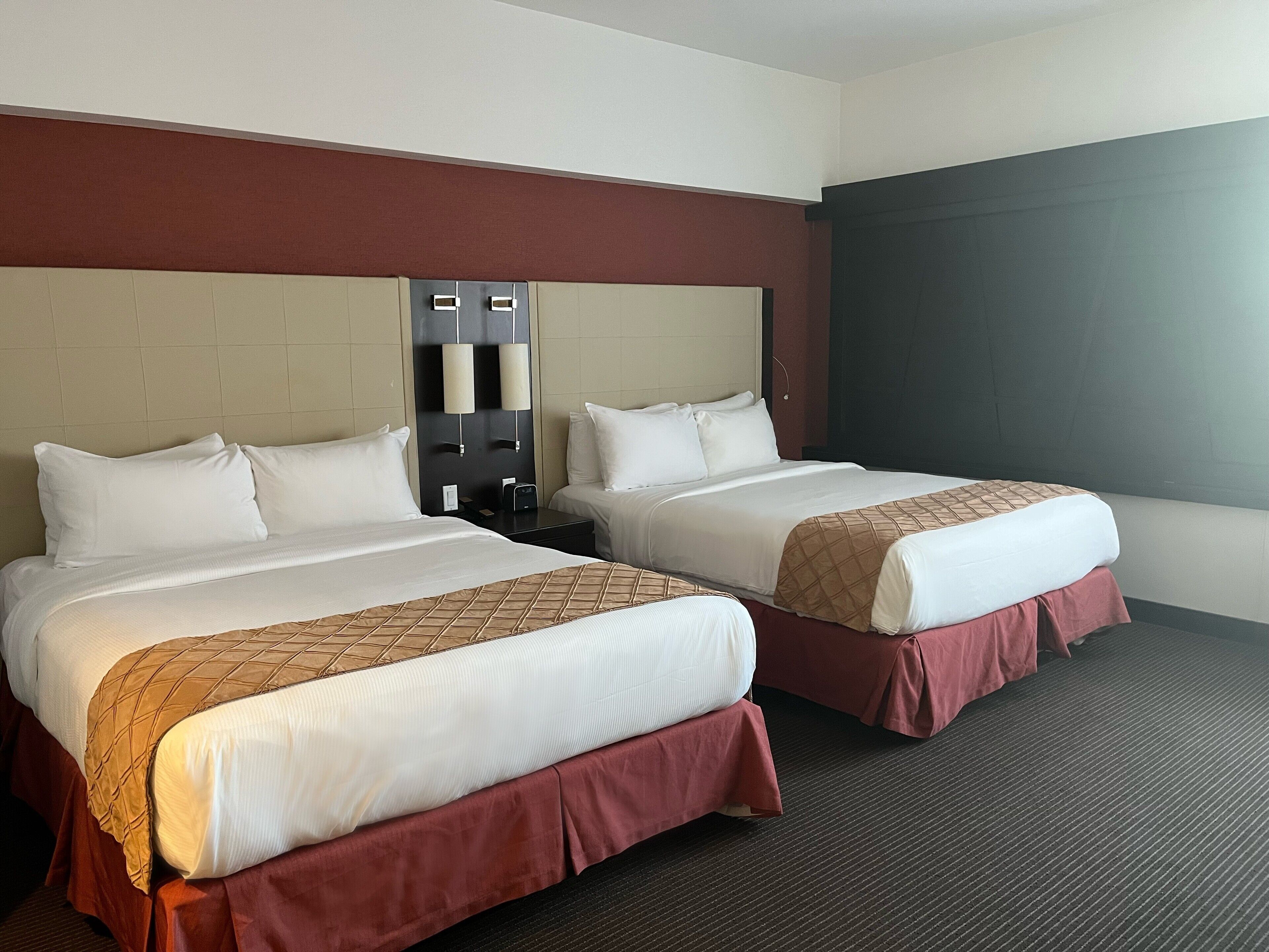 Habitación de hotel con dos camas, cada una con sábanas blancas y una colcha marrón. Tiene un cabecero de color oscuro y un gran ventanal que proporciona luz natural.