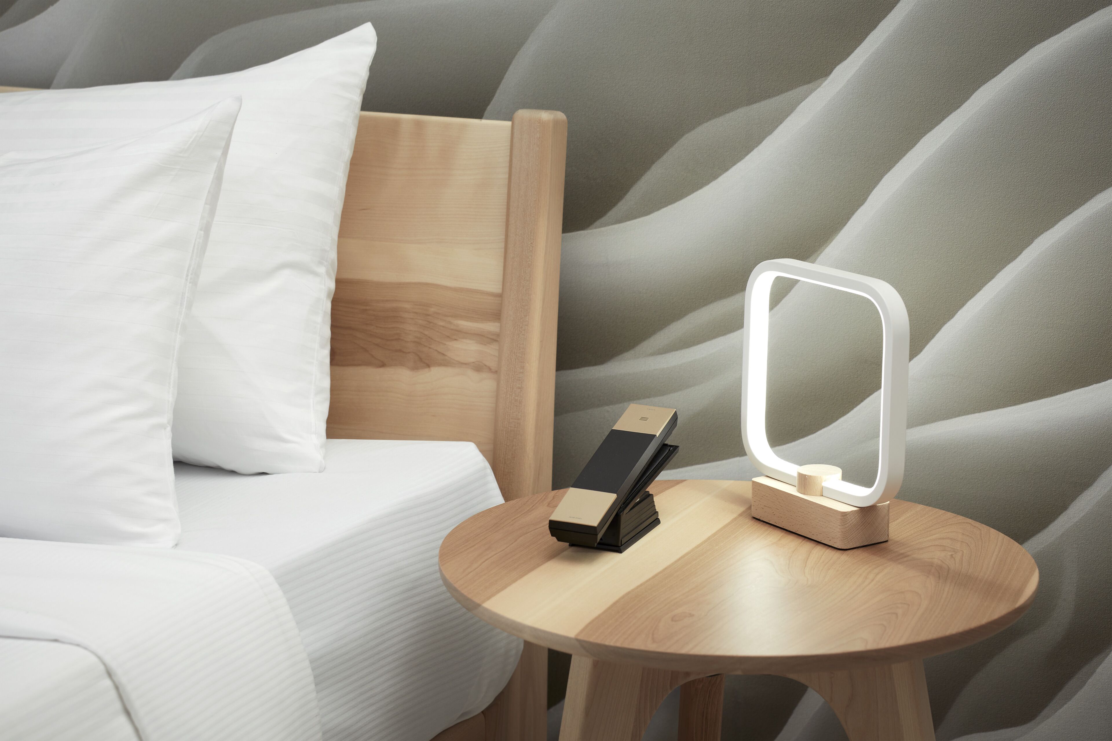 Una cama cuidadosamente hecha con ropa de cama blanca, un cabecero de madera y una mesita de noche con un espejo moderno iluminado y un objeto pequeño y rectangular encima.