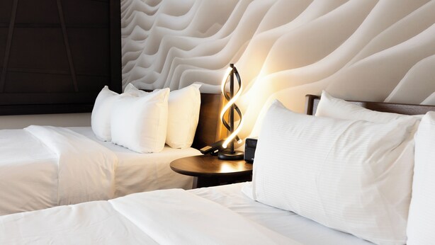 A hotel room with a modern design, featuring a large bed with white bedding and a unique textured headboard. There is a bedside table with a lamp and a small clock.