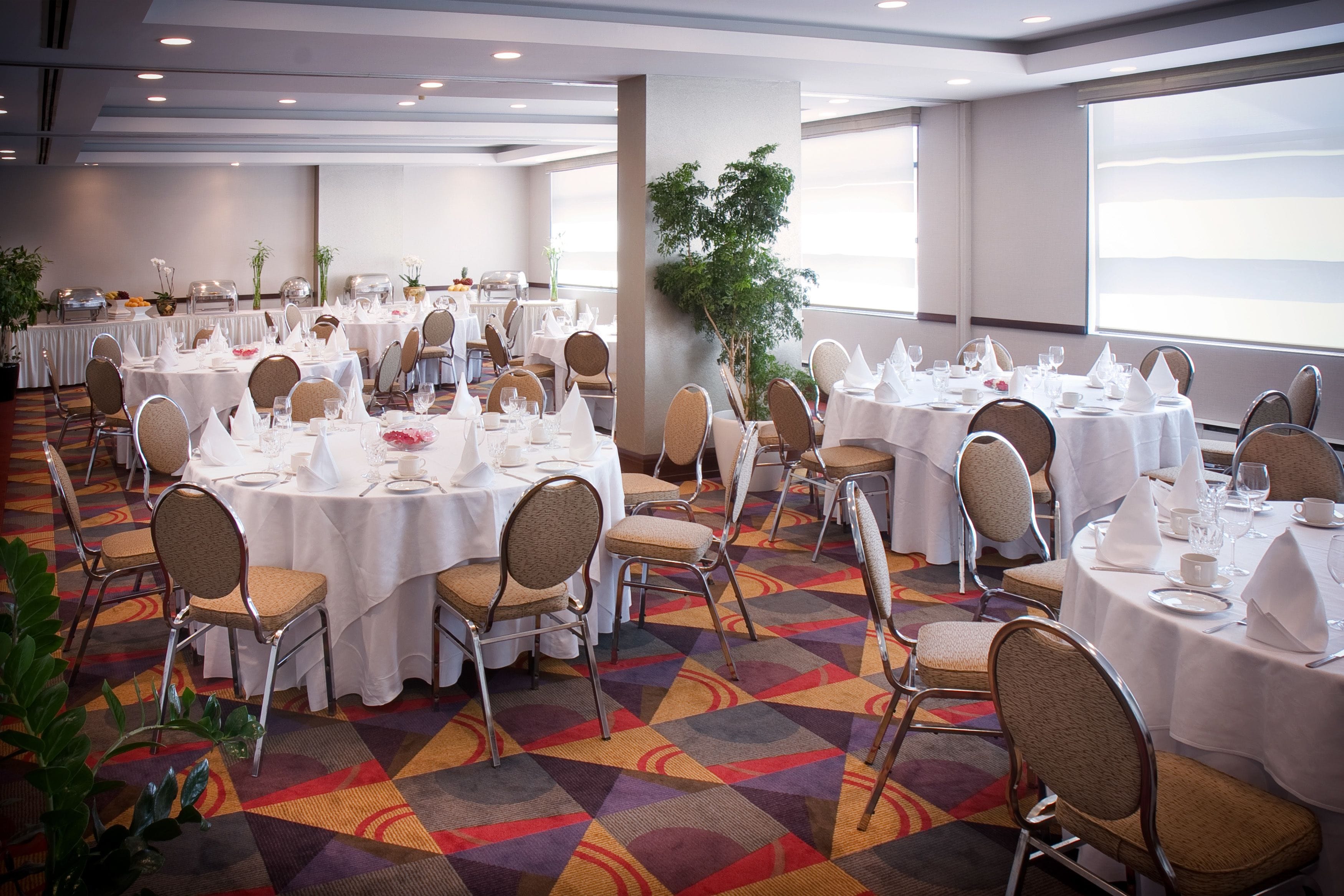 Un salón de banquetes con mesas redondas dispuestas para comer, sillas y un amplio ventanal que proporciona luz natural.
