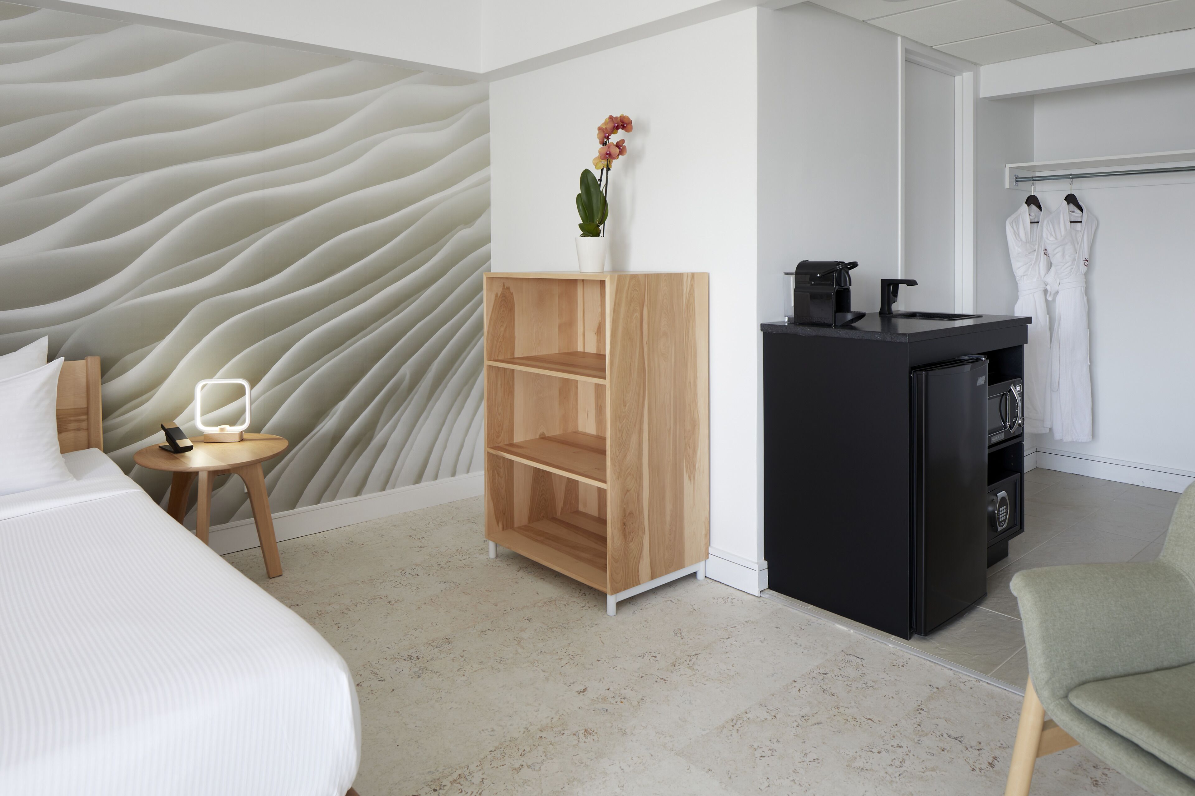 A hotel room with a bed, a wooden shelf with a potted plant, a black minibar, and a green chair.