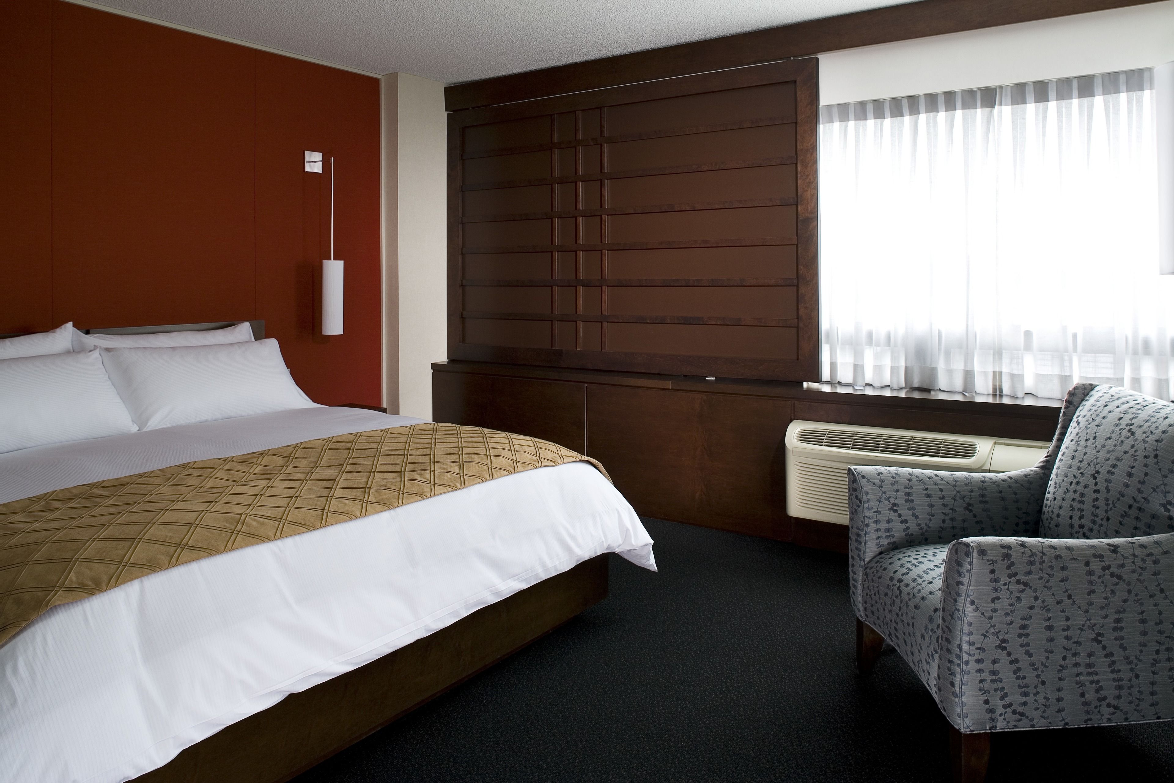 A hotel room with a large bed, a chair, a window with sheer curtains, and a wall-mounted air conditioner.