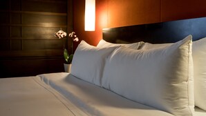 A neatly made bed with white linens, a potted plant, and a wall-mounted light fixture.