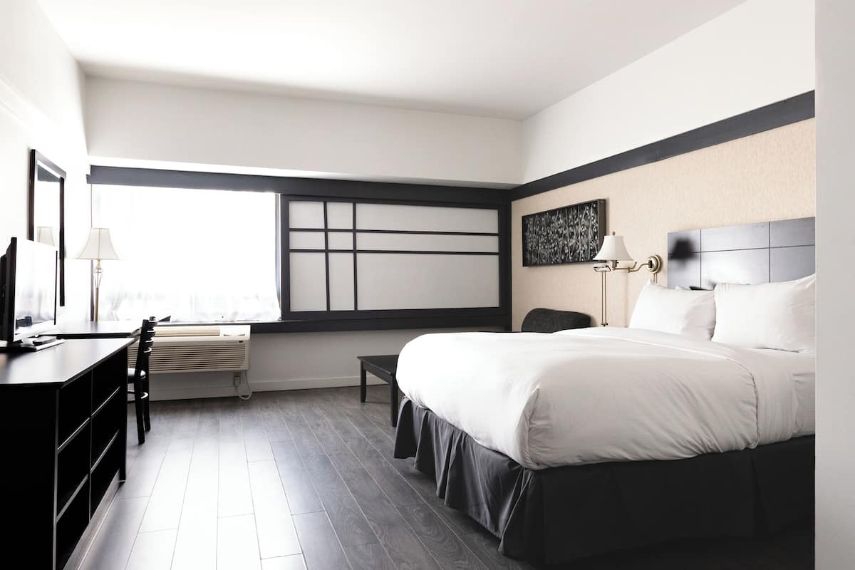 Une chambre d’hôtel moderne avec un grand lit, un bureau et une chaise. On y trouve une fenêtre avec des stores, un climatiseur fixé au mur et un tableau accroché au mur.