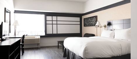 Una habitación de hotel moderna con una cama grande, un escritorio y una silla. Cuenta con una ventana con persianas, aire acondicionado de pared y un cuadro en la pared.