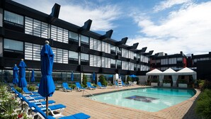 A pool area with lounge chairs and umbrellas, a pool, and a building with large windows and a modern design.