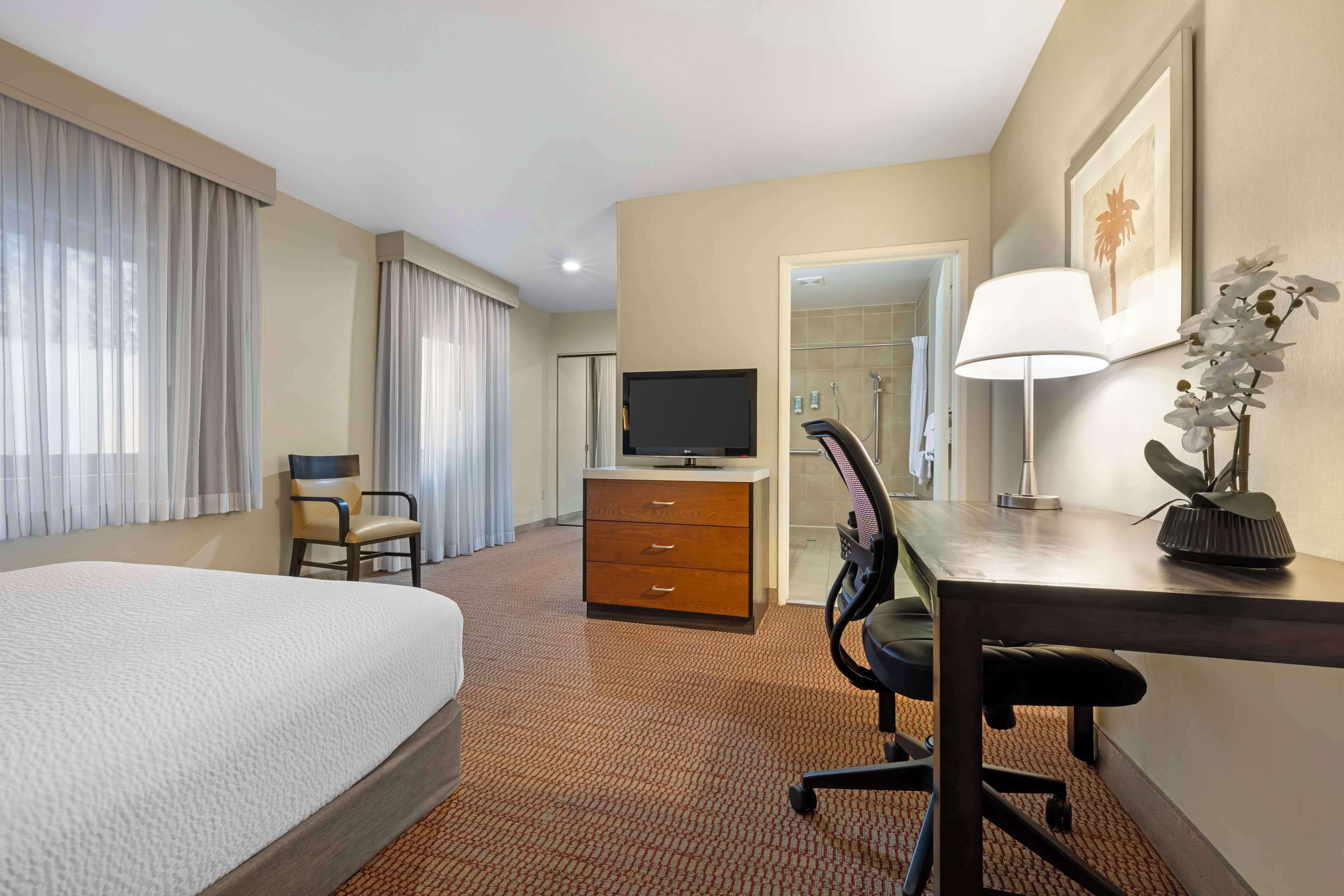A hotel room with a bed, a desk with a lamp, a television, a chair, a desk drawer, and a bathroom door.