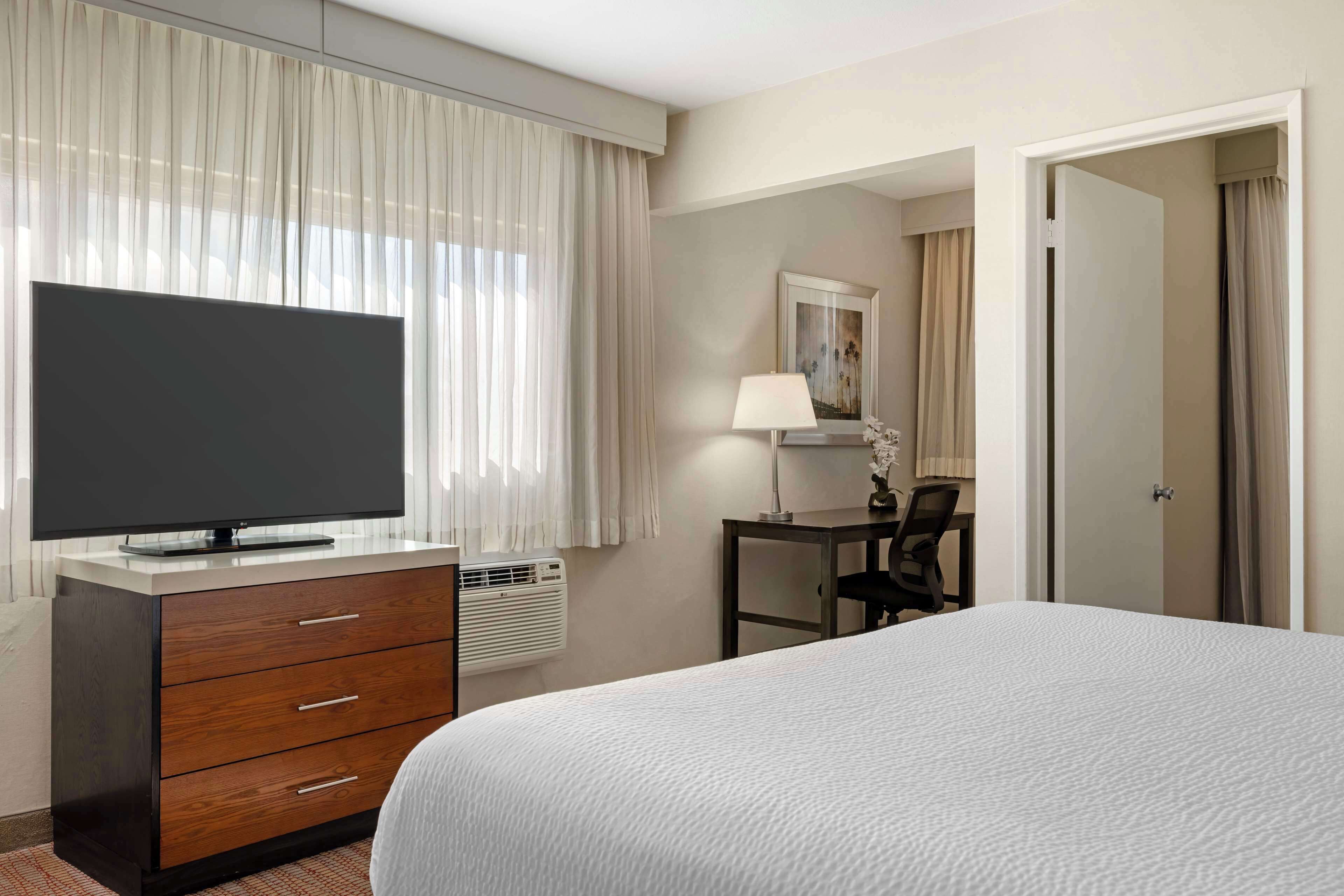 A hotel room with a bed, a TV on a dresser, a desk with a chair, and a door.