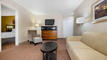 A hotel room with a beige sofa, a round coffee table, a TV on a wooden cabinet, a lamp, and a framed picture on the wall.