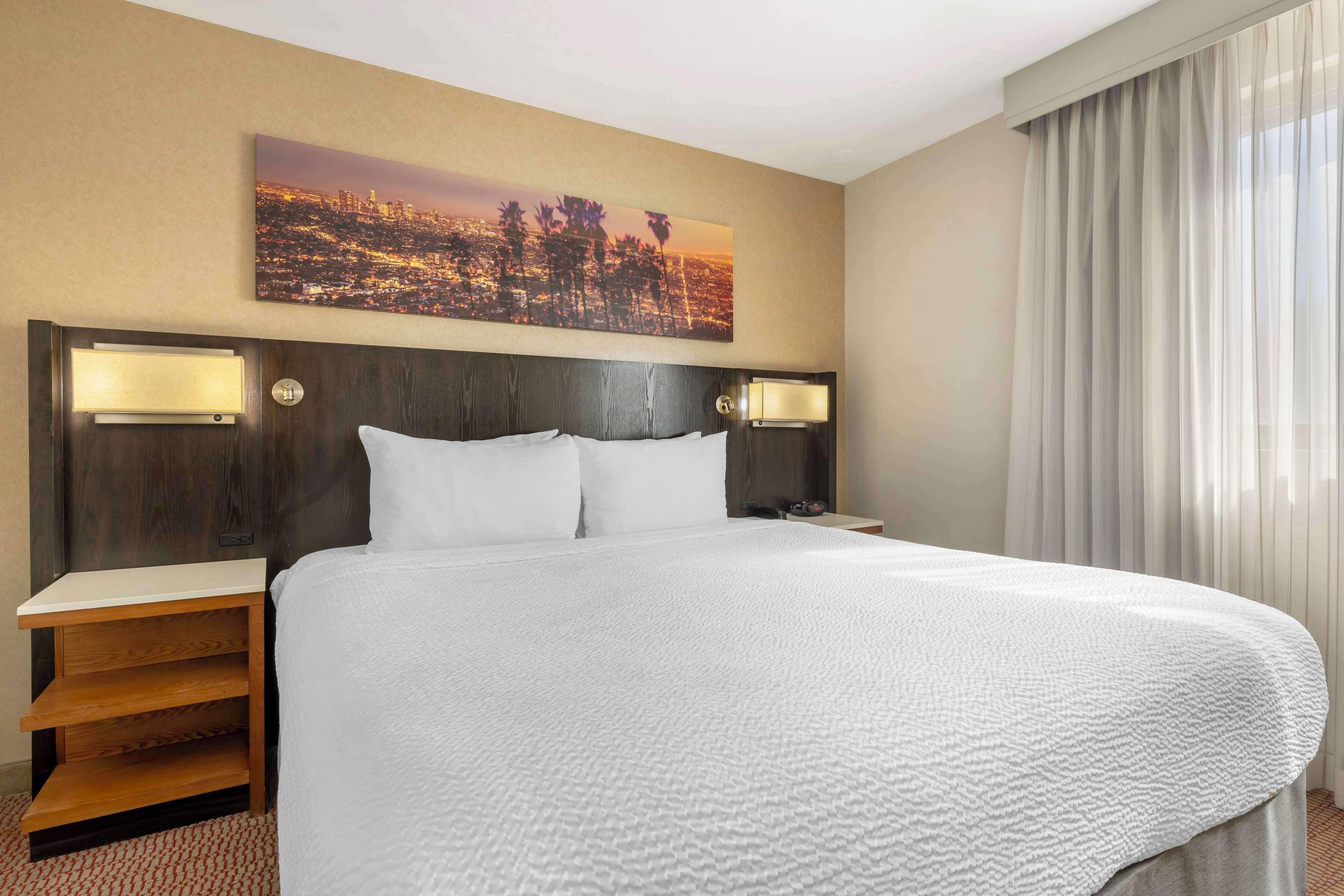 Habitación de hotel con una cama grande, dos mesitas de noche con lámparas, un cuadro de un paisaje urbano en la pared y una ventana con cortinas.