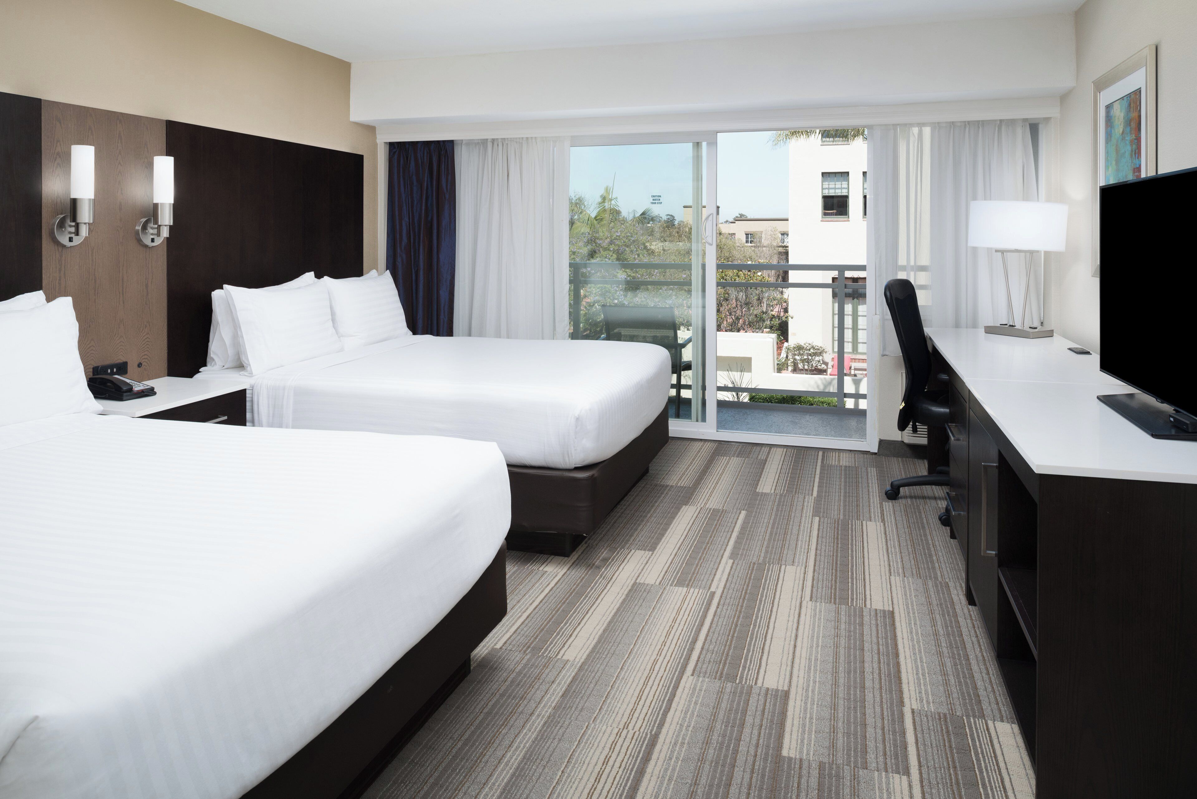 A hotel room with two beds, a desk, a chair, a TV, and a balcony with a view of the city.