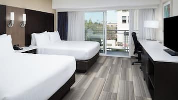 A hotel room with two beds, a desk, a chair, a TV, and a balcony with a view of the city.