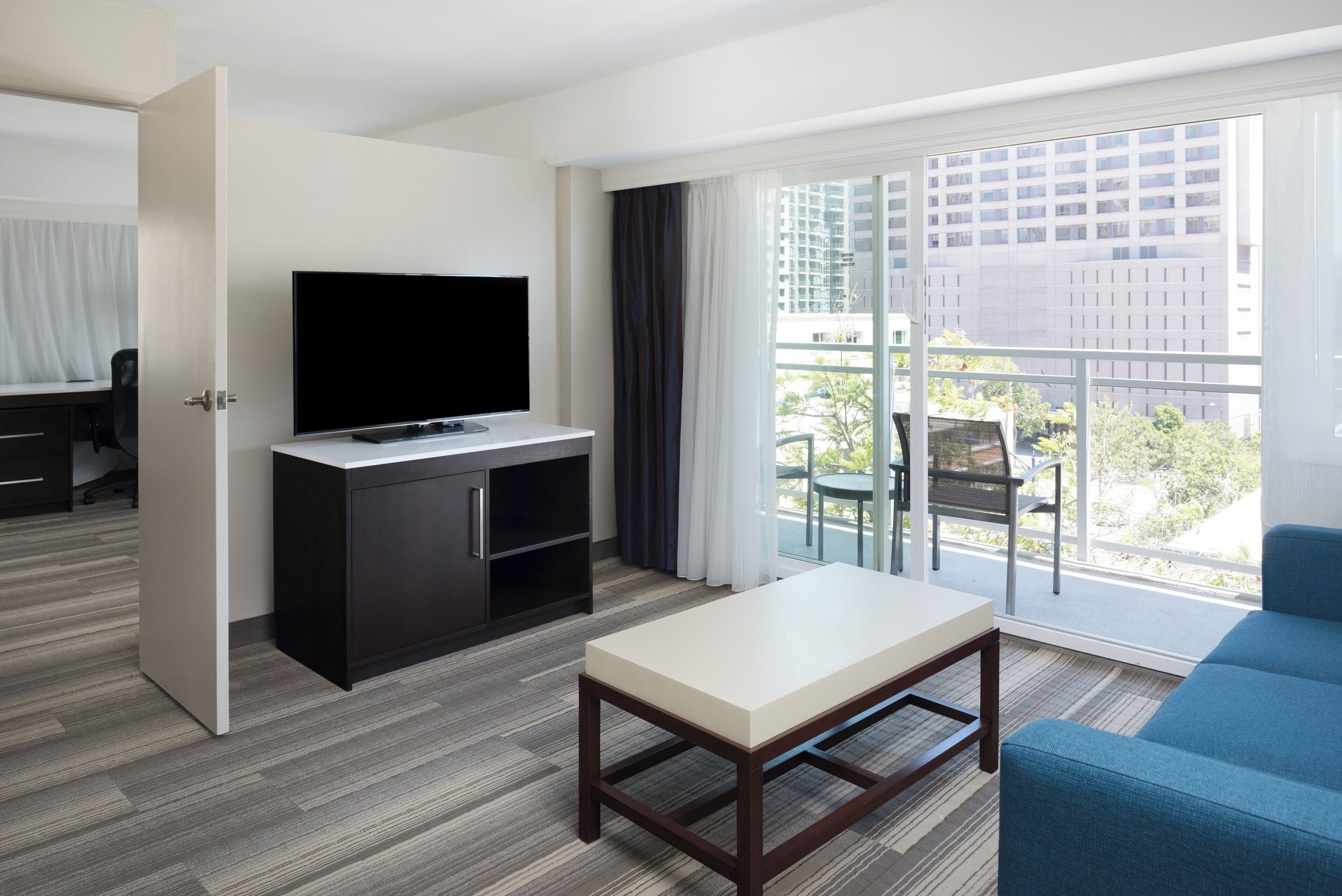 Una camera d'hotel con una televisione su un mobile, un tavolino, un divano blu e un balcone che si affaccia su edifici e alberi.